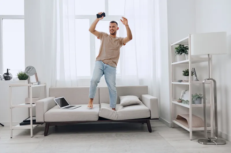 Joyful man celebrating freedom while stepping on the couch, embracing moments of digital detox and emotional minimalism in a bright, modern living space.