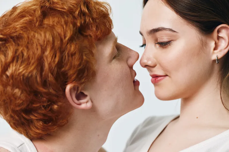Young couple with red hair and dark hair face each other closely with gentle expressions for love, intimacy and relationship on light background indoors.