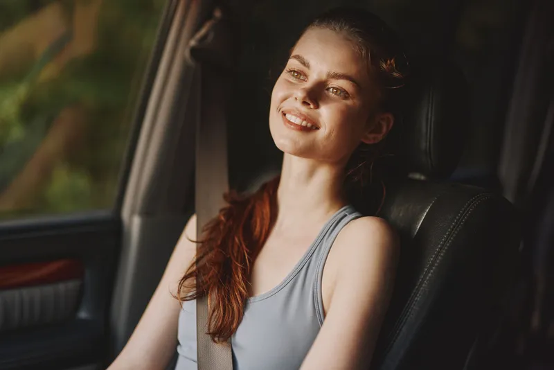 Smiling young woman with long hair in sporty attire, enjoying a sunny drive in a car, exuding happiness and relaxation, with a soft green background