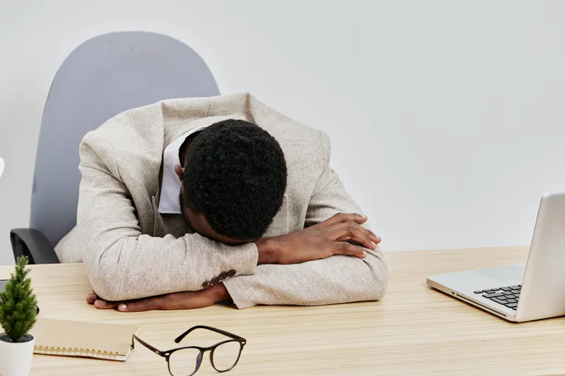 Tired young man with dark skin wearing beige sweater, resting his head on his arm at office desk beside laptop isolated on plain white background. People fatigue concept