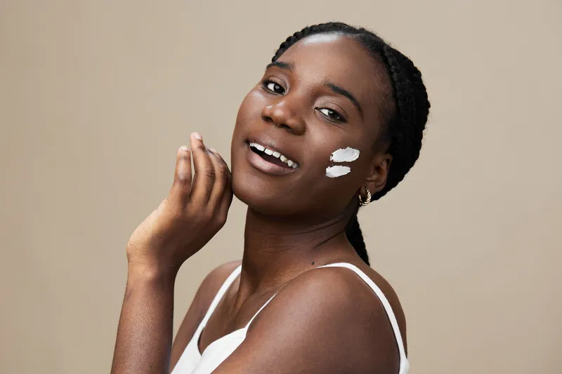Smiling young Black woman applying skincare cream on her face, showcasing a natural glow against a soft beige background, perfect for beauty and skincare concepts