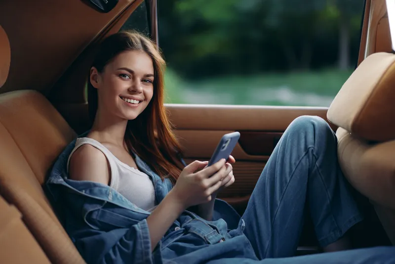 Young female model relaxing in a car, wearing denim outfit and smiling, showcasing a casual and joyful vibe surrounded by nature.