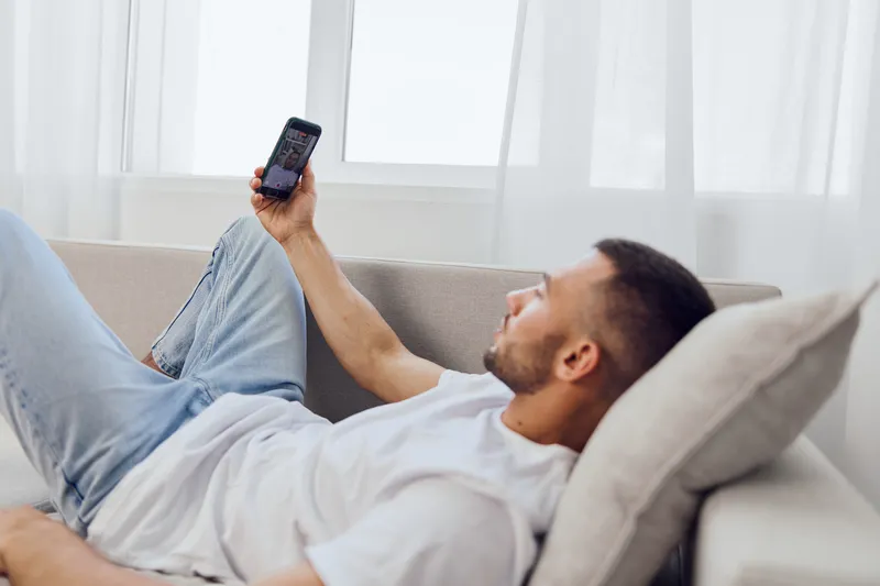 Relaxed man enjoying his smartphone while lounging on a couch in a bright room. Modern setting with natural light and a calm atmosphere evokes comfort and leisure.