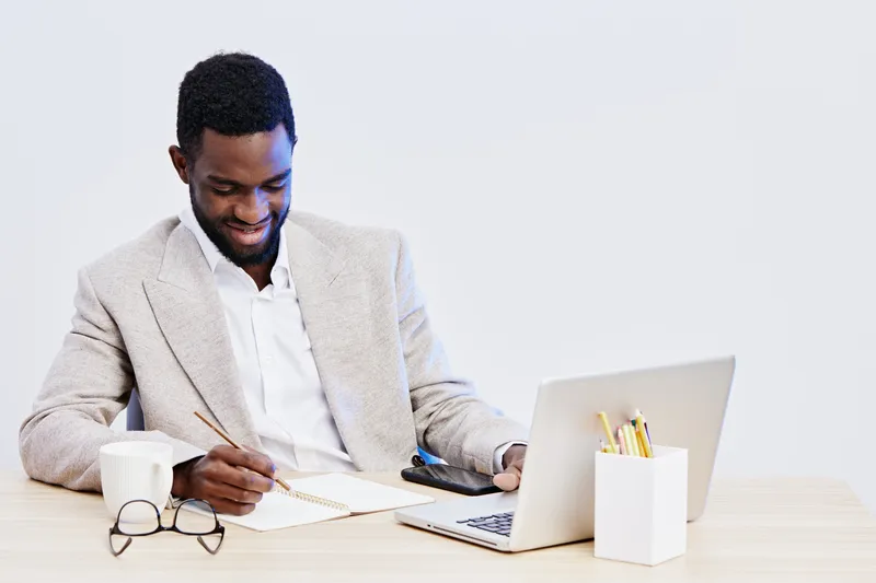 Happy young African-American man in beige blazer and white shirt working at desk with laptop, notebook, and glasses, looking at screen with smile, isolated on plain white background, studio shot. Peop