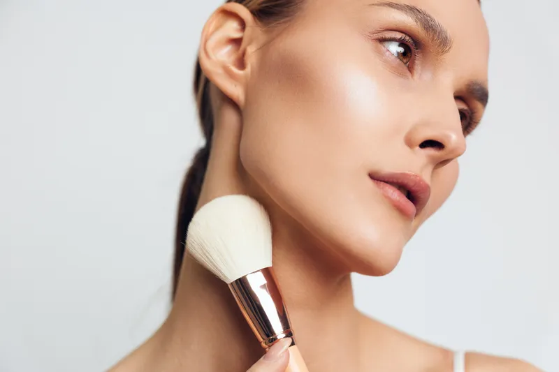 A young woman with a makeup brush applies powder to her cheek in a clean studio portrait, capturing a focused beauty routine, soft lighting, glowing skin, and a calm, intimate self-care moment.