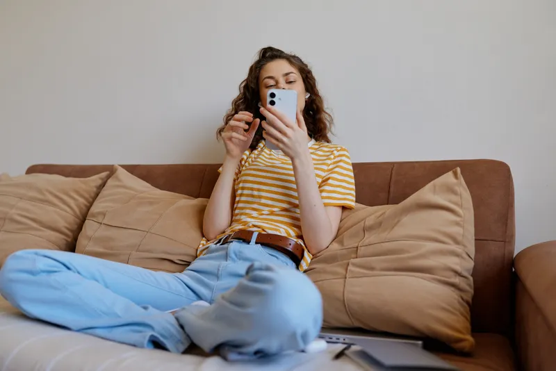 Young woman relaxing on a couch at home, looking at a smartphone. Casual lifestyle scene with warm lighting and a cozy, comfortable mood that suggests everyday self care and calm presence.