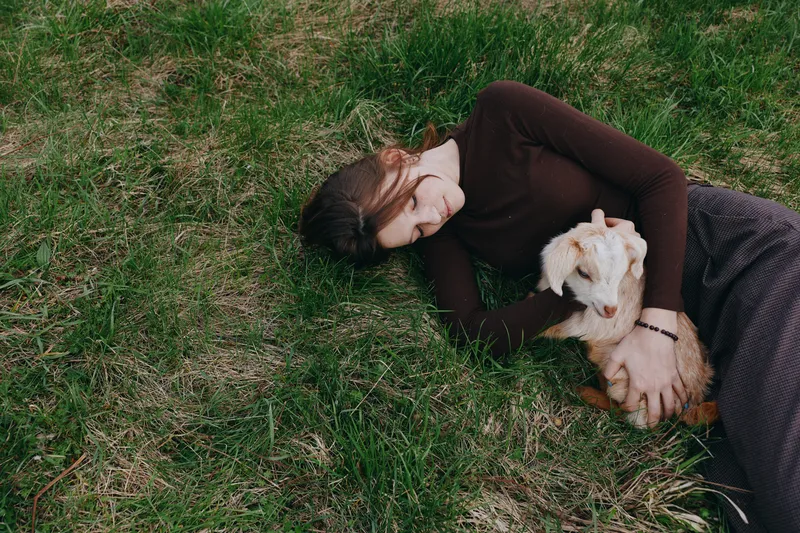 A tired woman lies on grass, embracing a small white dog in a sunlit outdoor setting. A moment of rest, companionship, and mental health through pet presence and gentle comfort.