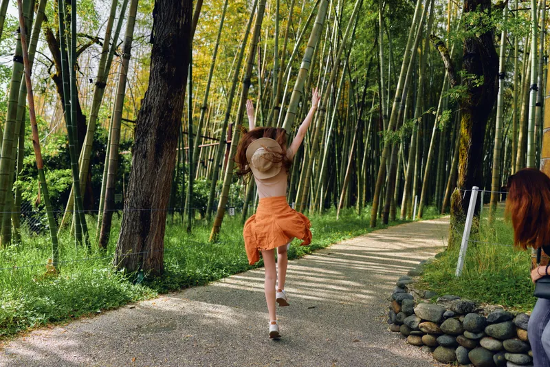 Bamboo woman running on gravel path in forest wearing hat and orange skirt, jumping with arms raised, back to camera in sunlit bamboo grove, joyful outdoor summer moment.