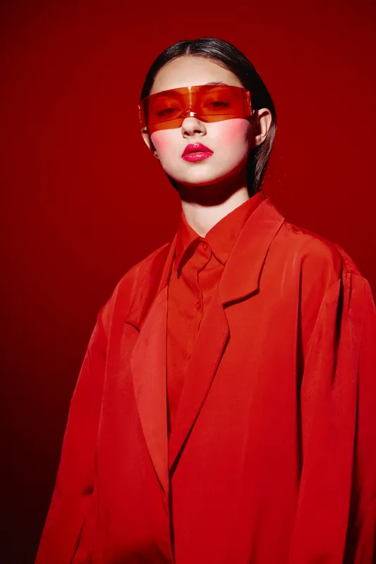 Fashion model in oversized red suit and futuristic orange sunglasses poses against a vivid red background. Skin glowing with dewy makeup adds bold artistic effect.