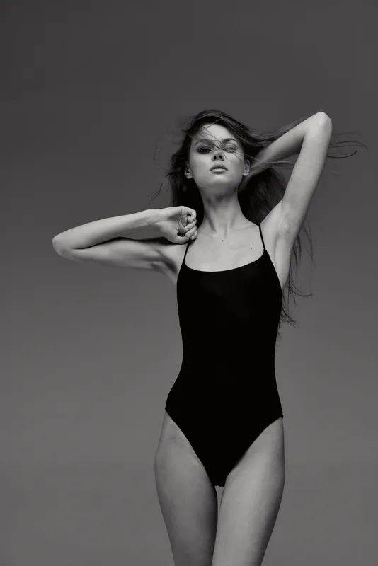 Black and white portrait of a young woman in a black swimsuit posing with confident expression against neutral background.