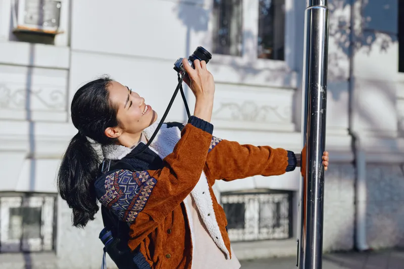 Photographer using a camera on a street, outdoor urban setting, leaning toward a pole in warm sunlight, a candid moment of creativity, exploration and artistic framing