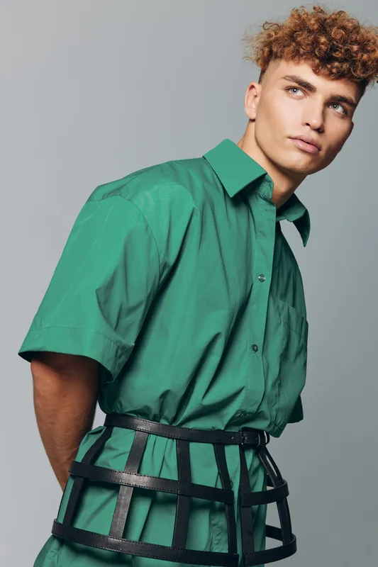 Young man with curly hair wearing green shirt and black cage belt posing against gray background in studio for fashion portrait and style concept.