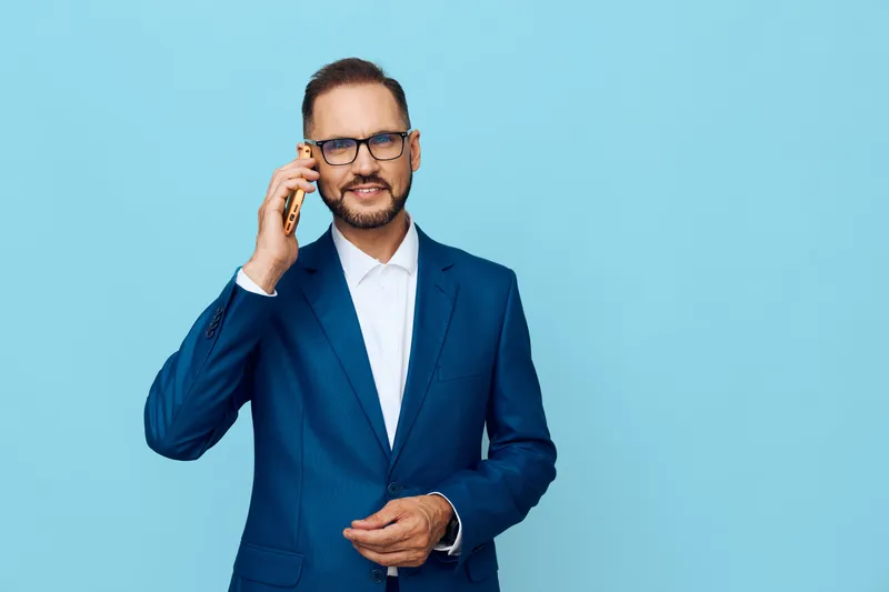 A confident business professional speaks on a mobile phone against a solid blue background, showcasing focus and composure as he discusses important work matters.