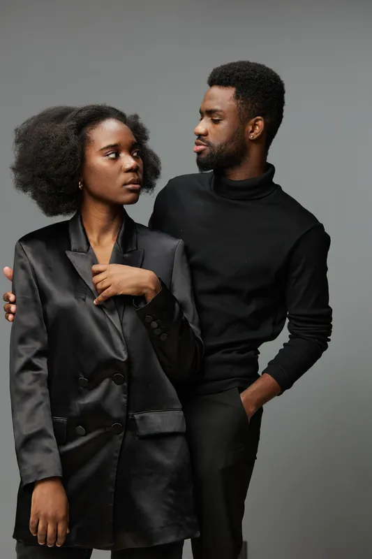 Elegant couple in black attire sharing a meaningful gaze against a neutral background, showcasing modern fashion and intimacy