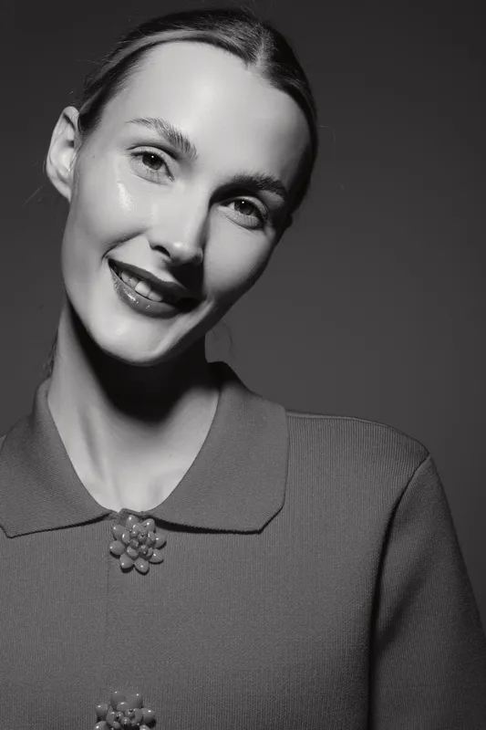 Portrait of a smiling Caucasian woman with light skin wearing a textured top and decorative brooch on dark background.