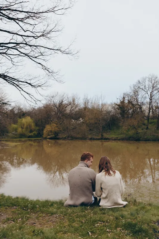 A young couple sits by a calm river in autumn wearing cozy sweaters while enjoying a quiet moment together in nature during a lifestyle outdoor scene.