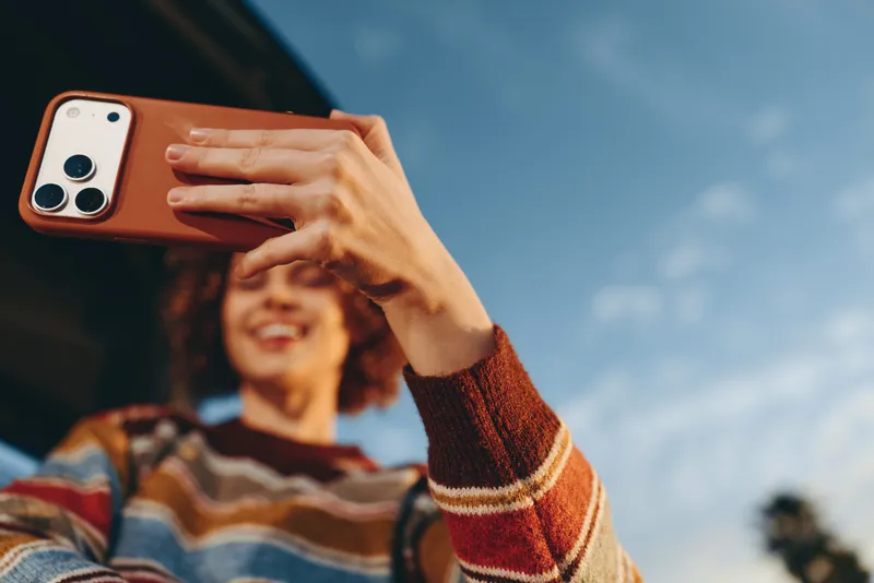 woman lifestyle outdoors, smiling and taking a selfie with smartphone in hand, warm sunshine and blue sky, relaxed posture, bright striped sweater, soft breeze, natural colors