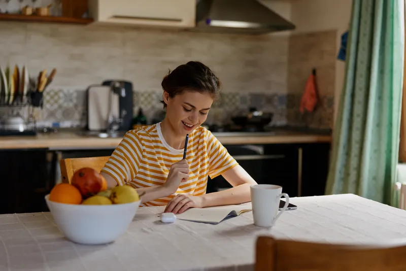 Cheerful woman at home, writing notes at a sunlit kitchen table, casual striped shirt, focused mood, soft breeze through window, warm colors, fruit bowl nearby, cozy lifestyle moment