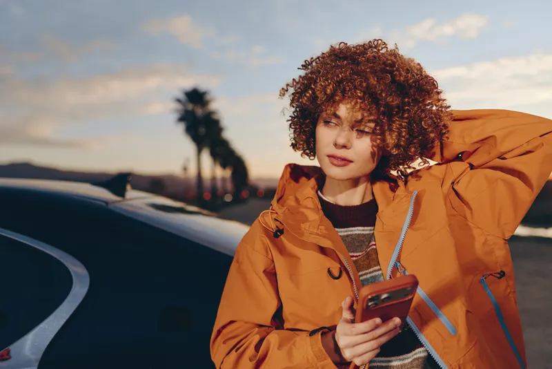 Lifestyle portrait of woman with curly hair in jacket outdoors at sunset, holding smartphone near car, relaxed smile, urban street road background, blurred palm trees