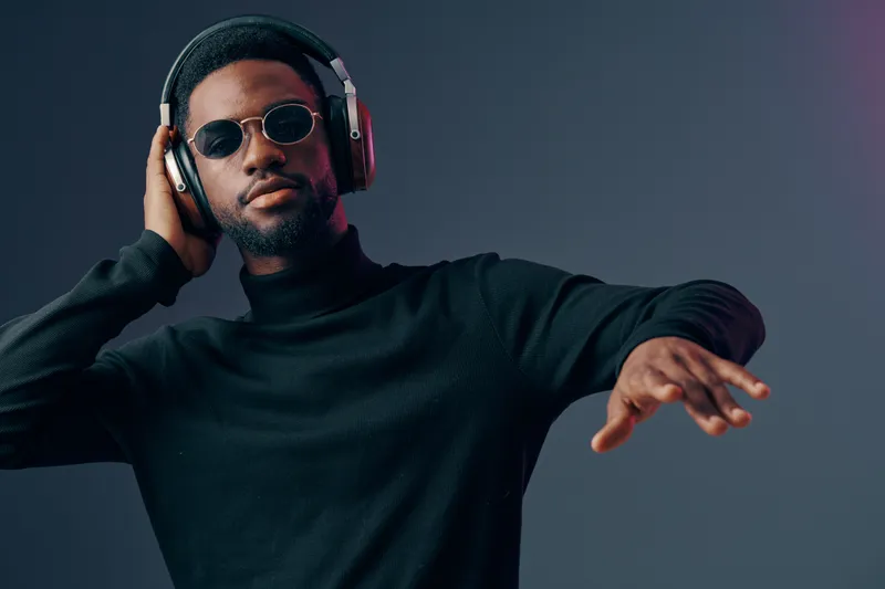 Portrait of a stylish Black man wearing headphones and sunglasses, enjoying music in a modern studio setting, featuring a cool color palette and a relaxed vibe