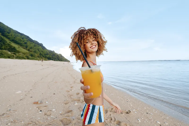 Smiling woman with curly hair on a beach, holding a juice drink with a straw. Summer vacation at the shoreline with sea and mountains behind, outdoors in warm sunlight, relaxed today.