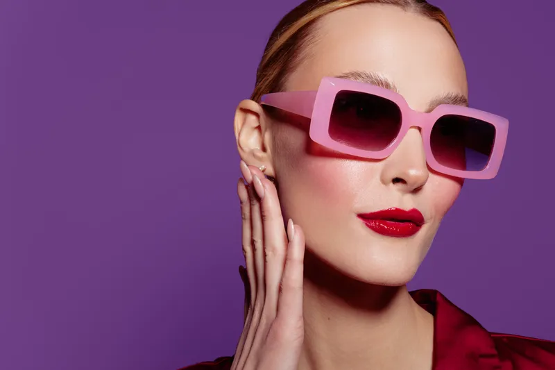 Stylish woman with pink sunglasses and red lipstick poses against purple background. Fashion portrait with trendy accessories and bold makeup. Concept of modern style and beauty.