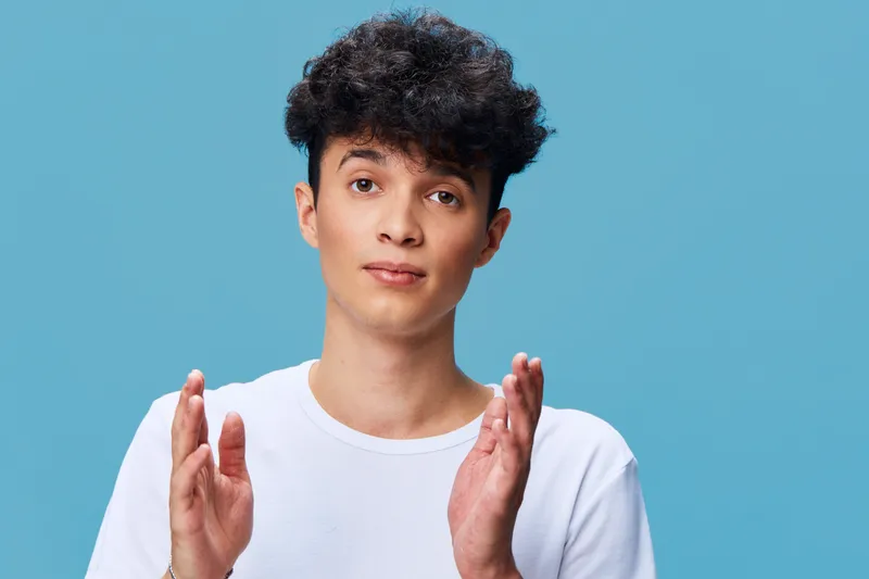 Young man in white shirt gesturing with hands against blue background, casual style, curly hair, calm expression, studio lighting.