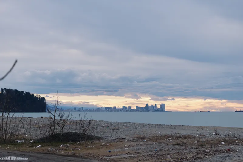 City skyline on the horizon above a quiet waterfront at dusk, with a rocky shoreline in the foreground and a cloudy sky reflecting soft light over calm waters.