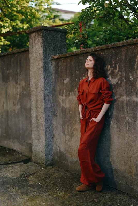 Woman in a red jumpsuit leaning against a textured outdoor wall, relaxed pose, soft sunlight, casual city vibe, thoughtful mood, natural tones