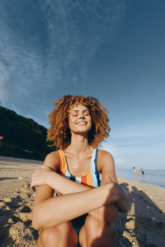 Woman at beach in sunlight, curly hair in yoga meditation pose, arms resting on knees on sandy shore, ocean horizon behind, calm relax concept, soft natural lighting