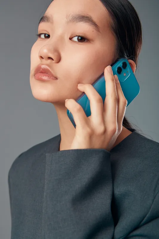Woman holding smartphone to ear with thoughtful expression wearing dark blazer on grey background. Closeup portrait of Asian female talking on mobile phone with modern device in hand.