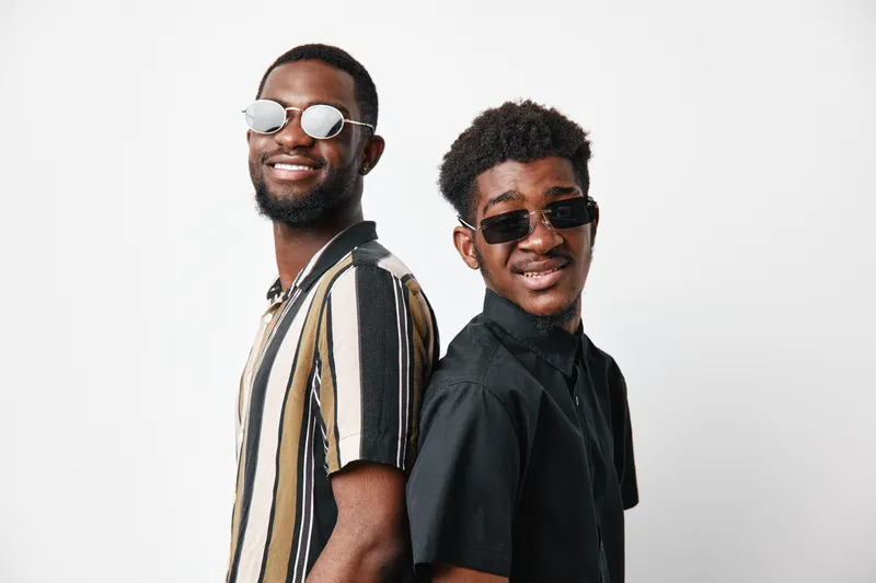 Two young black men with dark skin wearing sunglasses and casual shirts standing back to back smiling confidently against white background in studio portrait.
