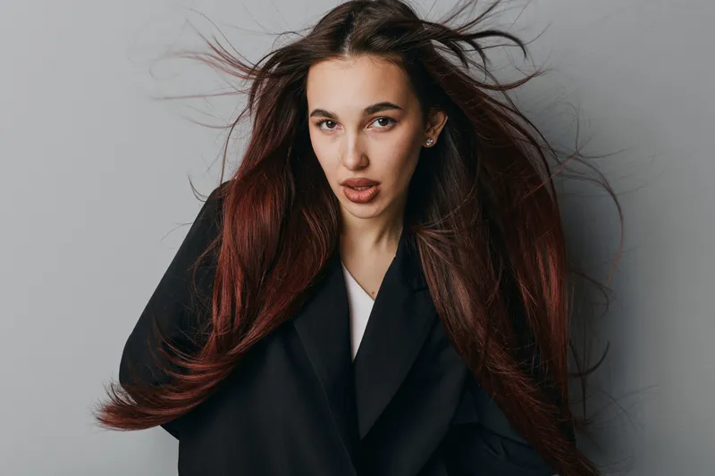 Woman with long brown hair wearing black coat posing against gray background with wind blowing hair for fashion, beauty, portrait and style concept in studio photography.