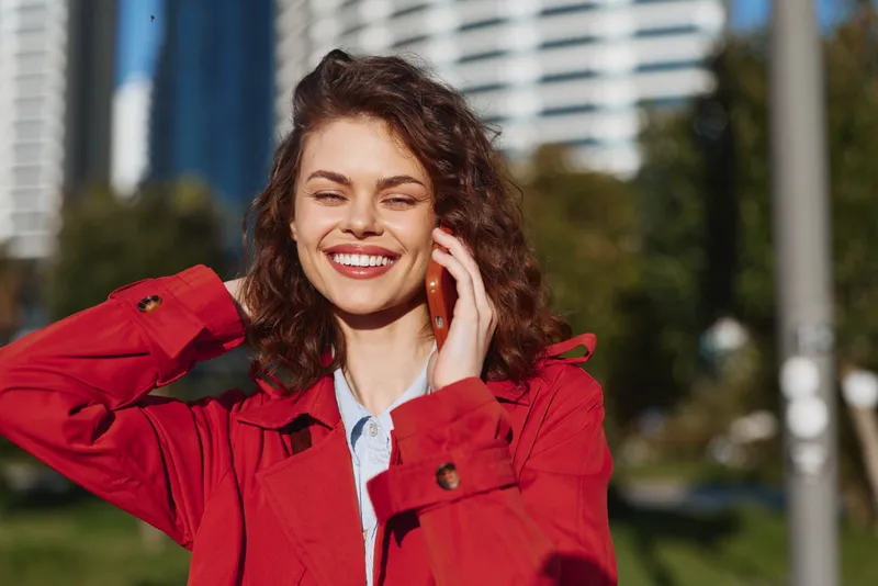 Young woman with curly brown hair in a red jacket chats on her phone outdoors, smiling under bright sunlight with urban buildings in the background during a casual daytime moment