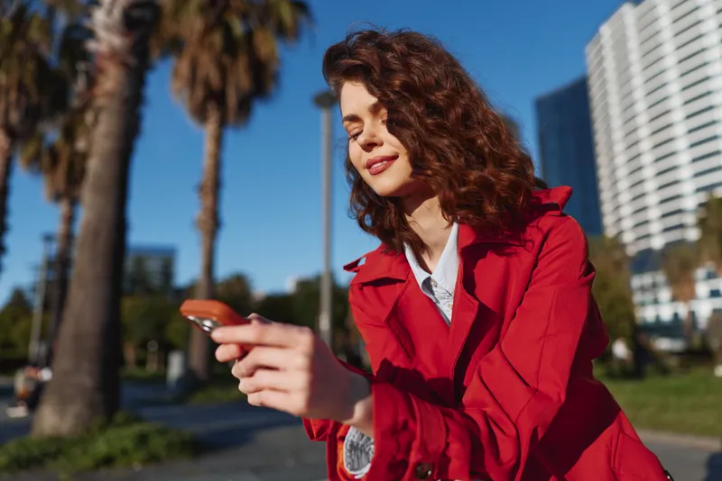 A stylish woman in a red coat uses a smartphone outdoors, sunny urban backdrop with palm trees and tall buildings, capturing casual street fashion and everyday tech.