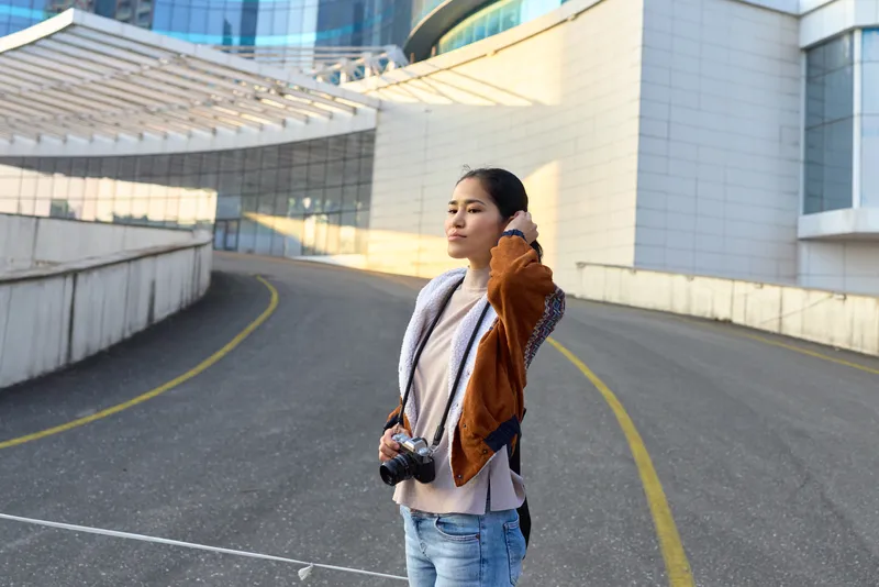 Woman photographer on a city street with a camera, casual jacket and jeans, exploring modern architecture, outdoor lighting, street vibe, travel inspired and candid urban moments