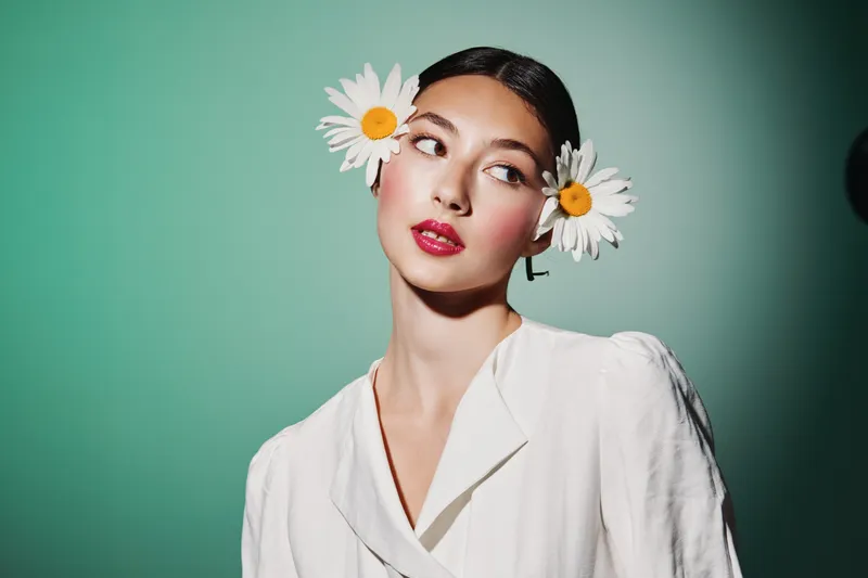 Young elegant woman with daisies in her hair posing against green background in white blouse. Concept of beauty, fashion, creative portrait, and floral style innovation.