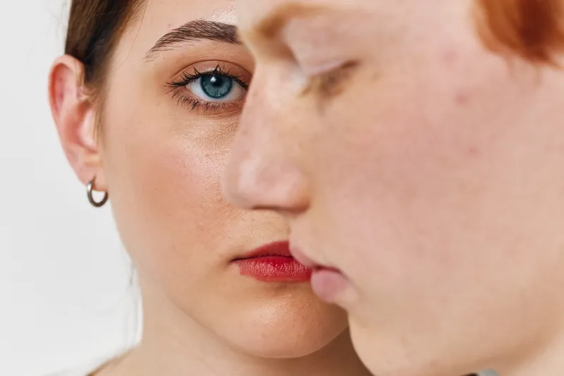 Woman and man with serious expressions in closeup portrait. Female face with blue eyes and red lips behind male profile, skin detail and emotion background in studio light.