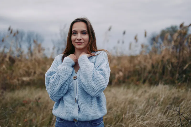 Natural portrait of a woman wearing a soft blue knit cardigan outdoors, conveying authenticity and credibility through real expression in calm autumn light and open field backdrop.
