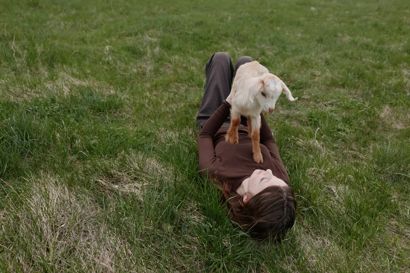 Lifestyle scene shows a woman lying on grass outdoors, cuddling a playful goat kid, conveying a tender moment of bond between human and animal in a rural setting.