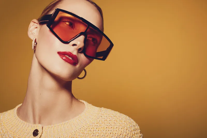 Stylish woman wearing oversized orange sunglasses and red lipstick poses against a warm yellow background. Fashion concept with bold accessories and confident expression.