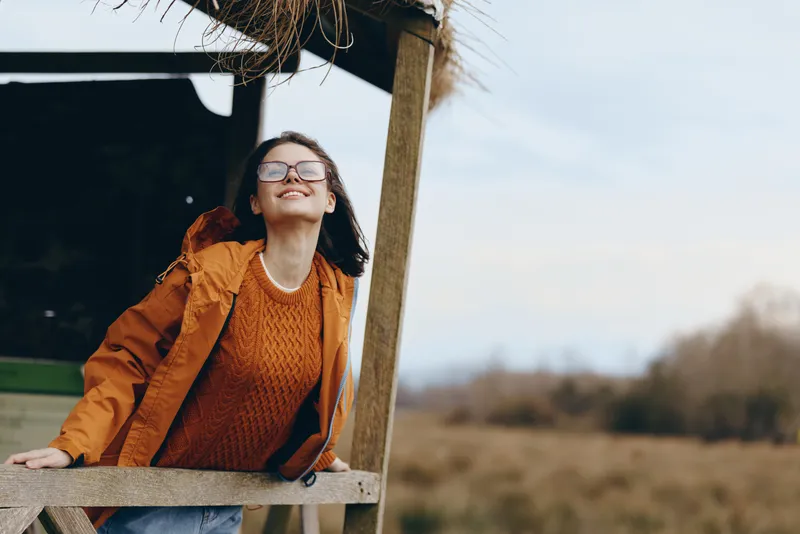 Woman lifestyle outdoors, smiling with glasses and hat, wearing orange jacket and knit sweater, leaning on wooden porch rail, sunlit countryside field, soft breeze, warm golden light