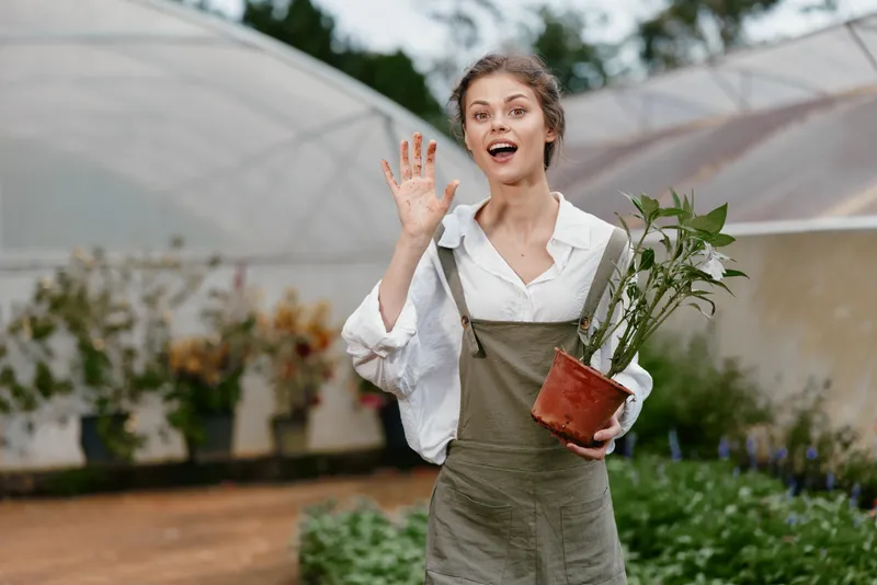 Young woman smiling and waving while holding a potted plant in a greenhouse, wearing a casual outfit, showcasing joy and connection to nature