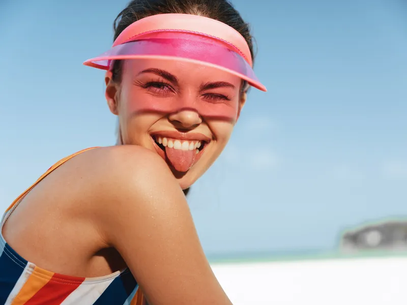Playful beach moment featuring a smiling woman wearing a pink visor, sunlit skin, and a colorful swimsuit. carefree expression captures energy, fun, and summer vibes.