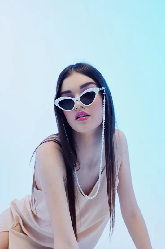 Young woman with long dark hair wearing white sunglasses and a beige top poses against a light blue background. Concept of woman beauty fashion and modern style.