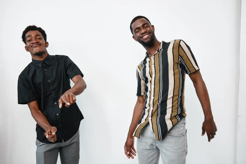 Two young black men with dark skin dancing and smiling together in casual clothes against white background. Friends enjoying music and having fun in relaxed atmosphere indoors.