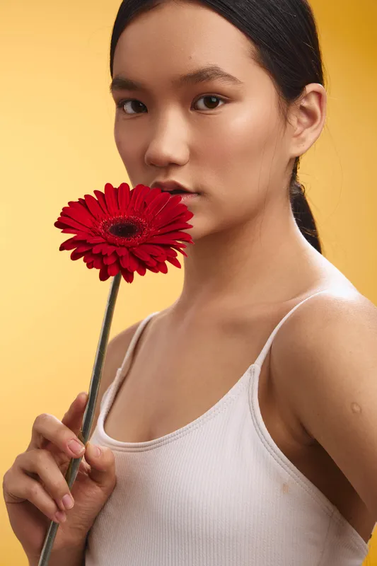 asian woman holds a bright red gerbera near her face, posing against a warm yellow backdrop, creating a soft studio portrait with subtle glamour and a calm, cheerful mood.