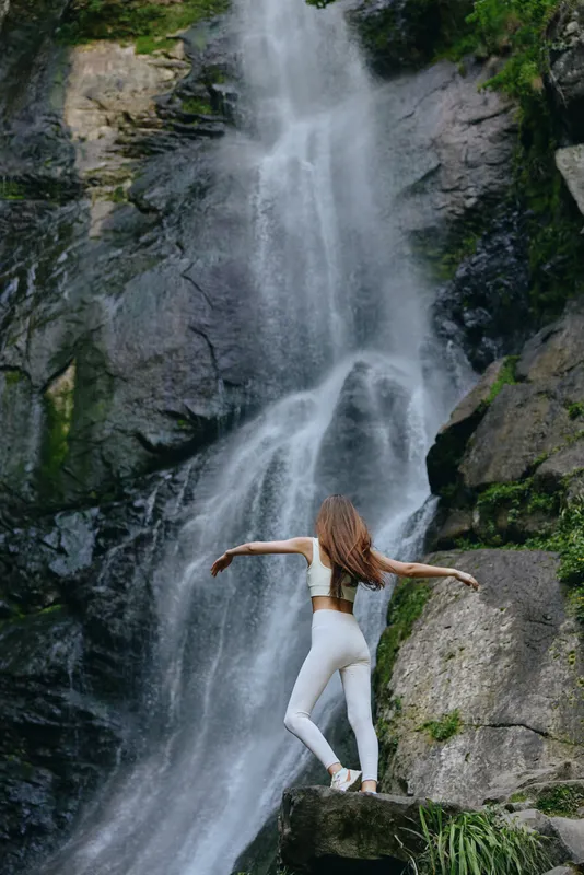 Woman in white activewear stands near powerful waterfall surrounded by rocks and lush greenery, enjoying nature and outdoor adventure with dynamic pose and flowing hair.