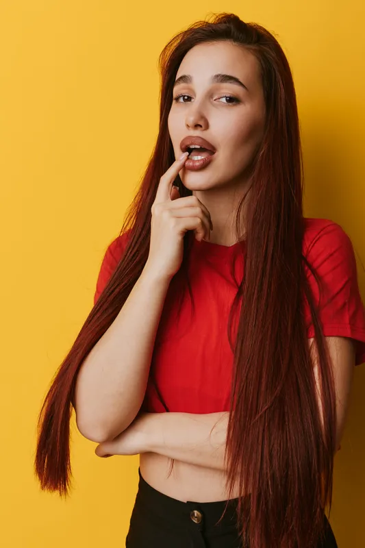 Young woman with long hair wearing red shirt posing with finger near lips on yellow background. Casual style, thoughtful expression and confident mood in studio portrait photography.