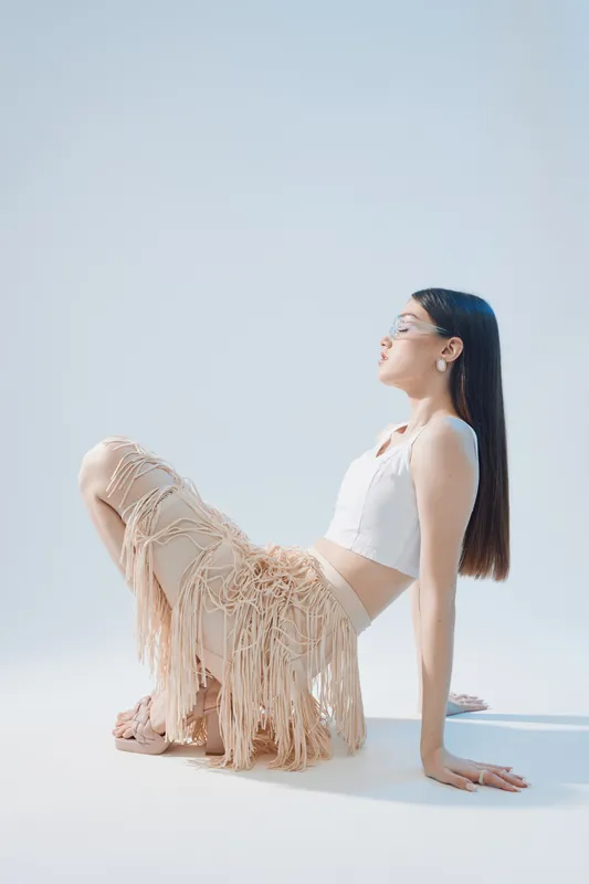 Young woman in stylish beige fringed pants and white crop top poses sitting on floor against light background. Concept for woman beauty fashion and modern style.